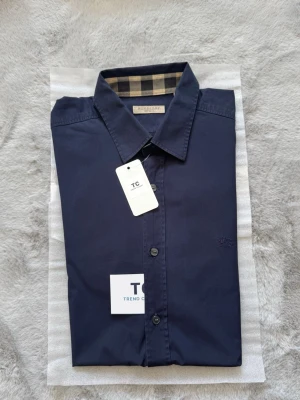 Burberry skjorta - Super fin marinblå burberry skjorta i storlek M, skjortan är i mycket fint skick utan några defekter. Skjortan är även äktighets verifierad. Burberry mönstret finns på båda ärmsluten och även runt om kargens indsida. Nypris 5000kr mitt pris endast 1200kr. Hör av er vid frågor. Mvh Trend closet🙌