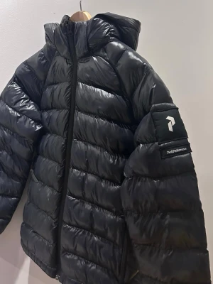 Svart dunjacka från Peak Performance - Snygg svart dunjacka från Peak Performance med huva och dragkedja framtill. Jackan har quiltad design, två sidofickor och logga på ärmen. Perfekt för kalla dagar och riktigt lätt att bära. Klassisk modell som funkar till det mesta.