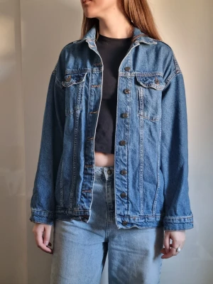 Oversized blå jeansjacka - Klassisk oversized jeansjacka i mellanblå denim med raka sömmar och bröstfickor med lock. Jackan har knappar framtill och vid ärmslut samt snygga kontrastsömmar. Perfekt att slänga över en t-shirt för en avslappnad look.