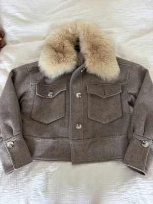 Kort beige pälsjacka med stor krage - Säljer en kort beige pälsjacka med stor fluffig ljus krage i fuskpäls. Jackan har två stora bröstfickor med lock och knappar framtill samt markerade muddar med knappar vid ärmsluten. Materialet är tjockt och har ett diskret fiskbensmönster. Använd och har missfärgning på pälsen uppe vid kragen men inget man påverkas av, sen två knappar som åkt av men tror jag har extra knappar som går att sy på. Är även villig att BYTA mot en SVART. Storlek XS/S