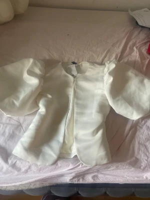 Vit blus med puffärmar - Trendig vit blus med stora puffärmar och rund halsringning. Blusen har en rak passform och är tillverkad i ett glansigt, lätt material som ger en elegant look. Perfekt för dig som vill sticka ut med en unik siluett.