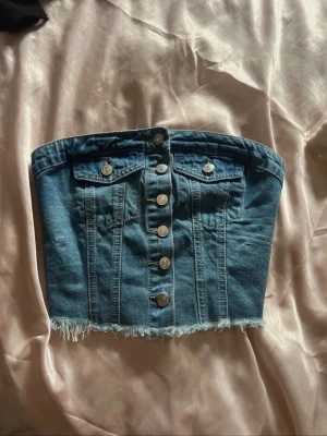 Blå jeans tubtopp med knappar - Säljer en blå jeans tubtopp med fransig nederkant och dekorativa silverknappar framtill. Toppen har två fickor med lock och knappdetaljer, samt markerade sömmar för en cool look. Perfekt för dig som vill sticka ut med denimstil.