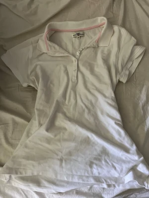 Vit pikétröja från Hampton Republic - Klassisk vit pikétröja från Hampton Republic med korta ärmar och krage med ljusrosa kant. Tröjan har knappar framtill och är gjord i bomull, perfekt för en avslappnad och stilren look.