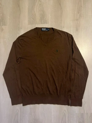 Brun V-ringad tröja Polo Ralph Lauren - Snygg brun V-ringad tröja från Polo Ralph Lauren med diskret broderad logga på bröstet. Tillverkad i mjukt material och har långa ärmar samt ribbade muddar vid ärmslut och nederkant. Perfekt för en stilren och klassisk look.