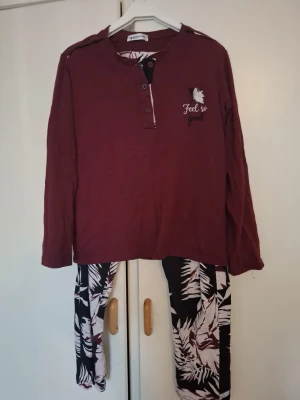 Vinröd pyjamas med bladmönstrade byxor - Snygg tvådelad pyjamas med vinröd långärmad tröja och trycket 'Feel so good' samt knappar vid halsen. Byxorna är svarta med stora vita och vinröda bladmönster. Mjuk och skön bomullskänsla, perfekt för chillkvällar hemma.