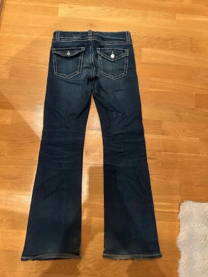 Mörkblå bootcut jeans med fickdetaljer - Snygga mörkblå bootcut storlek 158 jeans med kontrastsömmar och två bakfickor med lock och knapp. Jeansen har en klassisk femficksdesign och är tillverkade i denim med lätt stretch för skön passform.