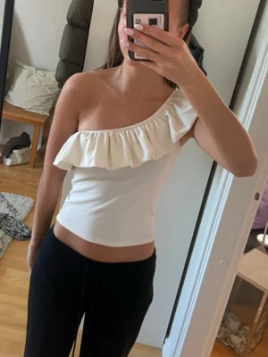 Vit one shoulder topp med volang - Säljer en vit one shoulder topp med bred volang längs halsringningen. Toppen är figurnära och har en snygg asymmetrisk design som ger en trendig look. Perfekt att matcha med både jeans och kjol för en cool stil.