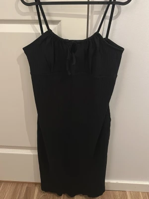 Svart axelbandsklänning med knyt - En svart klänning med smala axelband och knyt i halsringningen. Klänningen har en enkel, rak passform och är ärmlös. Perfekt för sommardagar eller när du vill ha en stilren look.