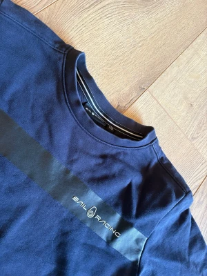 Mörkblå Sail Racing sweatshirt - Mörkblå sweatshirt från Sail Racing med en svart rand över bröstet och logga tryckt på vänster sida. Tröjan har rund halsringning och långa ärmar. Tillverkad i mjukt bomullsmaterial som känns skönt mot huden.