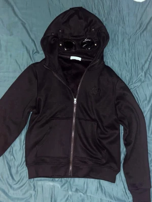 Svart hoodie med glasögon från C.P. Company - Säljer en svart hoodie från C.P. Company med dragkedja och ikoniska inbyggda glasögon i huvan. Hoodien har fickor framtill och broderad logga på bröstet. Perfekt för dig som vill sticka ut med en unik och streetinspirerad stil.