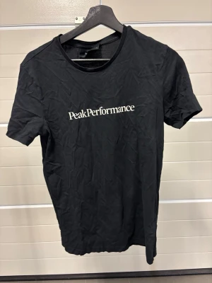 Svart Peak Performance t-shirt - Snygg svart t-shirt från Peak Performance med vit logga tryckt på bröstet. Klassisk passform och rund halsringning. Tillverkad i mjuk bomull som känns skön mot huden. Perfekt till jeans eller shorts för en clean look.