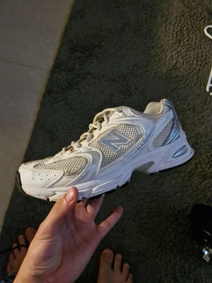 New Balance vita mesh sneakers - Snygga vita sneakers från New Balance med meshpaneler och silvriga detaljer. Skorna har en klassisk låg siluett, robust sula och reflekterande N-logga på sidan. Perfekta för dig som gillar sportig och clean stil.
