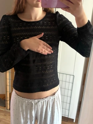 Svart spetsblus med lång ärm - Snygg svart blus i spets med lång ärm och rund hals. Blusen har ett genombrutet mönster och knapp i nacken. Perfekt för dig som gillar en feminin och trendig stil. 