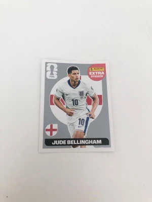 Panini FIFA 2026 Extra Sticker Jude Bellingham - Panini Extra Sticker med Jude Bellingham från FIFA World Cup 2026. Klistermärket har en silver bakgrund med Englands flagga och spelaren i vit matchtröja. Perfekt för samlare av fotbollskort och Panini-album. Kortet är självklart äkta. Köpt på pressbyrån. Kortet har ett litet veck en går fortfarande att klistra fast.