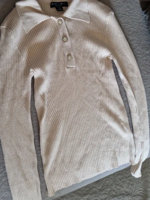 Beige ribbstickad tröja från Amisu - Snygg beige ribbstickad tröja från Amisu med lång ärm och stor krage. Tröjan har tre dekorativa knappar framtill med glittriga detaljer. Perfekt till jeans eller kjol för en trendig look.