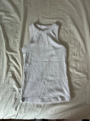 Vit ribbad tanktop - Säljer en klassisk vit ribbad tanktop i stretchigt material. Modellen är ärmlös med rund halsringning och har en tight passform. Perfekt att styla till jeans eller shorts för en enkel och clean look.