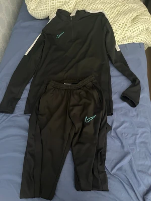Svart Nike träningsset med zip - Säljer ett svart träningsset från Nike med långärmad zip-tröja och matchande byxor. Setet har vita detaljer på ärmarna och gröna Nike-loggor på både tröja och byxor. Perfekt för träning eller chill. Materialet är mjukt och stretchigt för maximal komfort.