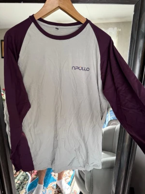 Vit och lila långärmad topp - Vit långärmad topp med lila ärmar och krage. Klassisk basebollstil med tryckt logga på bröstet. Perfekt för en avslappnad och sportig look. Tillverkad i mjukt material som känns skönt mot huden.