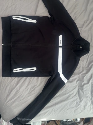 Hugo Boss tröja med krage - Snygg svart ziptröja från BOSS med vita dragkedjor på fickorna och en vit rand över bröstet. Tröjan har hög krage och raka ärmar, perfekt för en sportig och stilren look. Materialet känns mjukt och bekvämt. Storlek S men passar mig 186cm 78kg.