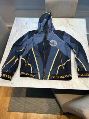 Blå och gul vindjacka med huva - Snygg vindjacka i blått och svart med gula detaljer och grafiskt mönster längs muddar och fåll. Jackan har huva, dragkedja framtill, två fickor med dragkedja och ett emblem på bröstet. Tillverkad i lätt, vindtätt material.