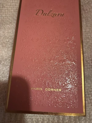Dulzura parfym från Paris Corner 5 ml  - Lyxig parfymflaska från Paris Corner med namnet Dulzura. Flaskan har en elegant design med guldmönstrad överdel och rosa glas. Förpackningen är rosa med gulddetaljer och vackert präglat mönster. Perfekt för dig som gillar exklusiva dofter och snygg inredning på sminkbordet.  5 ml flaska 