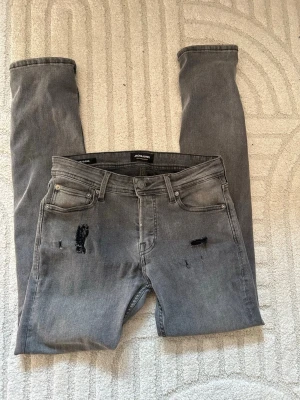 Grå slitna jeans - Säljer ett par grå jeans från Jack & Jones med slitna detaljer framtill. Jeansen har fem fickor, normal passform och klassisk knappgylf. Perfekta för en avslappnad och trendig look. Storlek W28/L32
