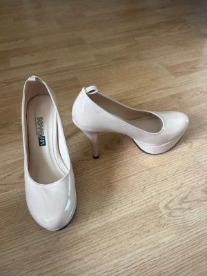 Beige lackpumps med röd sula - Snygga beige pumps i lack från Seven med rund tå och hög klack. Skorna har en cool röd sula och en klassisk siluett som ger extra höjd. Perfekta för dig som vill sticka ut lite extra med dina skor.