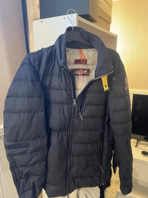Svart dunjacka från Parajumpers S - Snygg svart dunjacka från Parajumpers i storlek S. Jackan är superlätt, har dragkedja framtill, hög krage och klassiska Parajumpers-detaljer som gul rem och patch på ärmen. Perfekt för kalla vinterdagar och riktigt stilren look.