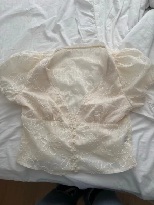 kräm vit blommig spetsblus med puffärm - Söt kräm vit blus med blommig spets och puffiga korta ärmar. Djup v-ringning och knappar framtill ger en romantisk vibe. Perfekt till jeans eller kjol för en trendig look. Materialet är tunt och lätt, vilket gör den luftig och bekväm.
