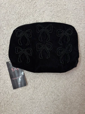Svart sammetsnecessär med rosetter - Söt svart necessär i mjuk sammet från Hunkemöller. Framsidan är dekorerad med sex broderade rosetter i pärlformade detaljer. Perfekt för smink eller småprylar. Dragkedja upptill och stilren design. Aldrig använd. Prislapp kvar, nypris 279kr.