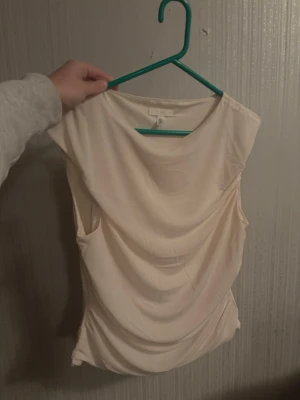Vit ärmlös topp från H&M - Säljer en stilren vit ärmlös topp från H&M. Toppen har draperad front och rund halsringning, vilket ger en snygg och modern look. Perfekt att matcha med jeans eller kjol för en trendig outfit.