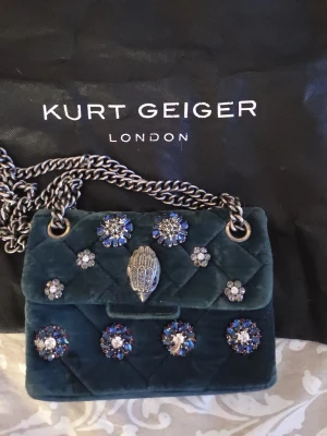 Kurt Geiger väska  - Superfin mörkgrön Kurt Geiger väska som tyvär inte används längre. Jag har endast använt den några få gånger så den är i fint skick förutom en liten fläck och en kristall som har lossnat, men det märks inte när man bär den! Pris kan diskuteras så kom gärna med prisförslag!🤍