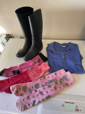 Ridkläder med blå piké och knästrumpor - Säljer ett kit med ridkläder: svart höga ridstövlar storlek 30, blå pikétröja från CRW i storlek XS samt tre par ridstrumpor i olika rosa nyanser och mönster. Perfekt för dig som gillar hästar och vill ha färgglada detaljer i stallet.