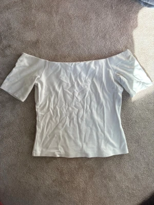 Vit offshoulder topp i bomull - Snygg vit offshoulder topp i mjuk bomull. Toppen har korta ärmar och en figurnära passform som ger en clean och stilren look. Perfekt att matcha med jeans eller kjol för en trendig outfit.