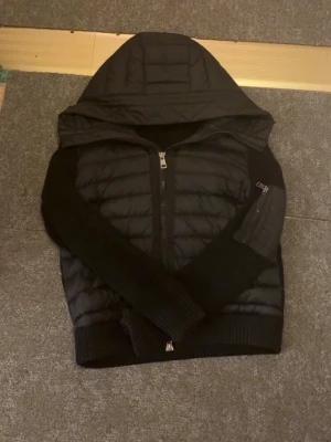 Svart pufferjacka med huva - Säljer en svart pufferjacka med huva och dragkedja framtill. Jackan har stickade ärmar och muddar samt en ficka med dragkedja på ena ärmen. Kan byta jacka eller diskutera pris vid snabb köp 
