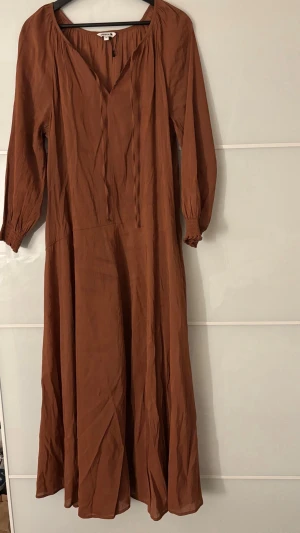 Brun långklänning med v-ringning - Säljer en snygg brun långklänning med v-ringad hals och långa ärmar. Klänningen har en avslappnad passform och är tillverkad i ett lätt material som ger ett fint fall. Perfekt för dig som gillar enkel men stilren design.