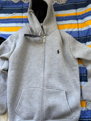 Grå zip hoodie från Ralph Lauren - Snygg grå hoodie med dragkedja från Ralph Lauren. Klassisk design med svart logga på bröstet, stor huva och två rymliga fickor framtill. Perfekt för chill dagar och enkel att matcha med allt. Mjuk bomullskänsla och riktigt skön passform.
