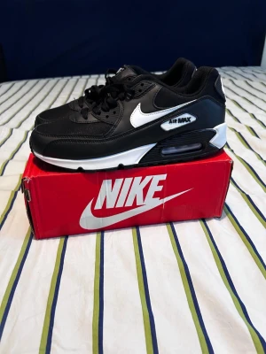 Nike Air Max 90 svart/vit sneakers - Säljer ett par klassiska Nike Air Max 90 sneakers i svart med vita detaljer och vit sula. Skorna har den ikoniska Air Max-dämpningen och tydlig Nike-logga på sidan. Perfekta för dig som vill ha en stilren och tidlös sneaker med skön passform.