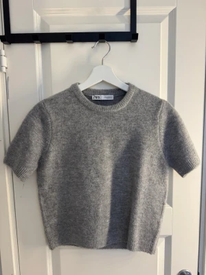 Grå stickad t-shirt från ZARA - Kortärmad stickad t-shirt i mjuk grå ullmix från ZARA. Tröjan har rund halsringning och ribbade kanter för en clean look. Perfekt att styla lager på lager eller bära som den är.