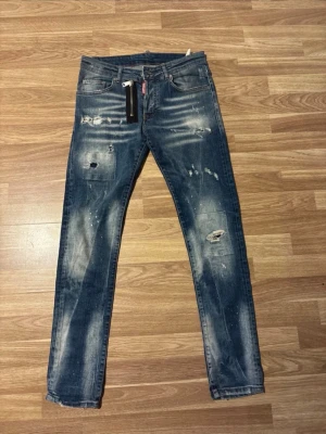 Blå slitna jeans från Dsquared2 - Säljer ett par blå jeans från Dsquared2 med slitningar, hål och färgstänk för en riktigt trendig look. Jeansen har smal passform, klassisk femficksdesign och dragkedjedetalj framtill. Perfekta för dig som gillar streetstyle och vill sticka ut.