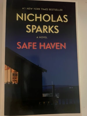 Safe Haven av Nicholas Sparks - En romantisk och spännande roman av Nicholas Sparks. Omslaget är mörkblått med gul och röd text, och visar ett hus vid vattnet i skymningen. Boken handlar om Katie som flyr sitt förflutna och hittar kärlek och trygghet i en liten stad. Perfekt för dig som gillar gripande kärlekshistorier.