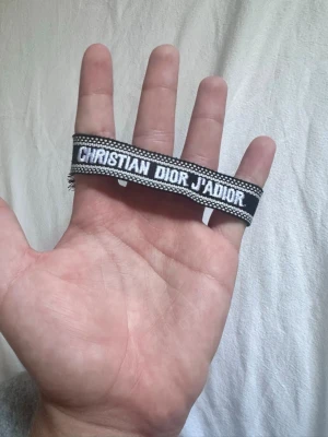 Christian Dior J'Adior tygarmband - Snyggt svart och vitt tygarmband från Christian Dior med texten 'J'Adior' broderad. Armbandet har justerbar knytning med tofsar i ändarna och en cool, trendig look som passar perfekt till din outfit.