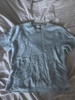 Ljusblå stickad t-shirt - En ljusblå stickad t-shirt med rund hals och korta ärmar. Tröjan har ribbade kanter vid hals, ärmslut och nederkant. Perfekt för en avslappnad och stilren look. Aldrig använd.