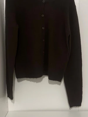 Mörkbrun basic kofta - En enkel och stilren mörkbrun kofta med knappar framtill. Koftan har lång ärm och rak passform, perfekt att slänga över en t-shirt eller topp. Materialet ser mjukt och lite fluffigt ut, vilket ger en mysig känsla.