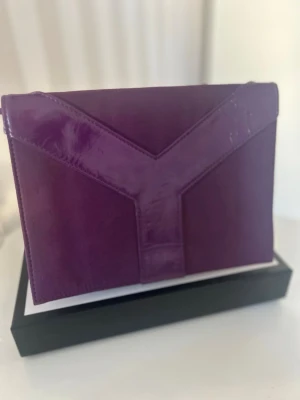YSL Kuvertväska - Säljer denna superfina och stilrena kuvertväska/pouch från Yves Saint Laurent i en djuplila färg.  Väskan har en lyxig design i mjukt tyg/mockaimitation med det ikoniska Y-mönstret i glansigt material på framsidan.   Perfekt som en elegant clutch till festen, som sminkväska eller för att organisera mindre tillhörigheter i en större reseväska.  Märke: Yves Saint Laurent  (GWP / Gift With Purchase)  Mycket bra skick med minimalt slitage. (Det glänser lite av kameran men inget slitage finns.