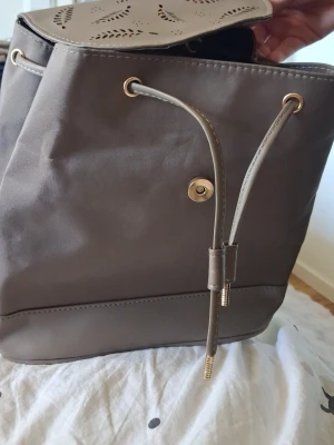 Grå ryggsäck med dragsko - Snygg grå ryggsäck i slätt konstläder med dragskostängning och guldfärgade metalldetaljer. Locket har ett dekorativt mönster och väskan har en stilren, modern look. Perfekt för dig som vill ha en enkel men trendig accessoar.