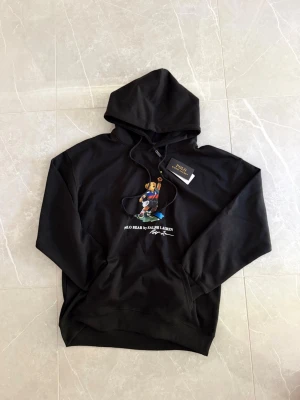 Svart hoodie Polo Bear Ralph Lauren - Svart hoodie från Ralph Lauren med Polo Bear-tryck framtill där björnen spelar tennis. Klassisk huva med dragsko och känguruficka. Mjuk bomullskänsla och ribbade muddar vid ärmslut och nederkant. Perfekt för en avslappnad och trendig stil.
