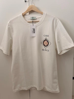 Vit Casa Blanca t-shirt med tryck - Säljer en stilren vit t-shirt från Casa Blanca med ett unikt grafiskt tryck och text på bröstet. T-shirten har rund hals och korta ärmar, perfekt för en clean och modern look. Materialet känns mjukt och skönt mot huden.