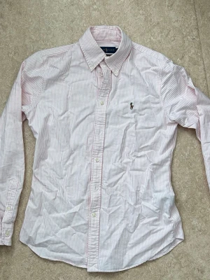 Rosa och vit randig skjorta från Ralph Lauren - Klassisk skjorta från Ralph Lauren i 100% bomull med rosa och vita smala ränder, långärmad och med knappar framtill. Skjortan har en broderad logga på bröstet och en snygg krage. Perfekt för en clean och stilren look. Storlek M men sitter mer som S eller mindre enligt mig