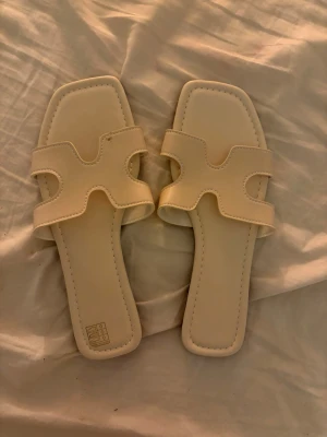 Beige slip-in sandaler med fyrkantig tå - Stilrena beige sandaler med öppen design och fyrkantig tå. De har breda remmar i syntetmaterial som korsar foten och ger en modern look. Perfekta för sommaren och lätta att matcha till olika outfits.